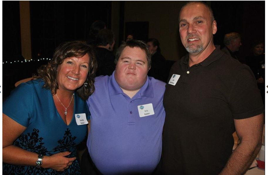 A cheerful group of three people posing for a photo at an event, with two adults flanking a young man in the middle, all wearing name tags and smiling faces.