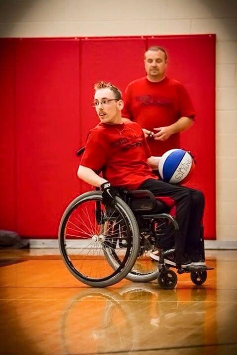 A focused wheelchair basketball player holding a ball, with a coach standing in the background, both wearing matching red team shirts in a gymnasium.