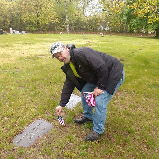 A person placing a small american flag next to a grave marker in a green cemetery.