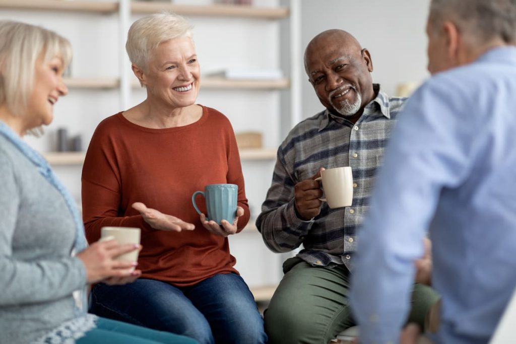 Group of senior citizens in a group. They are having a discussion and are smiling. Some have coffee mugs in their hands.